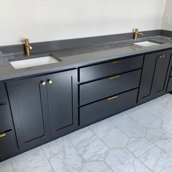 A modern bathroom vanity with dark cabinets, gold handles, and two sinks. The countertop is gray, and the flooring features white hexagonal tiles. The faucets have a sleek gold finish.