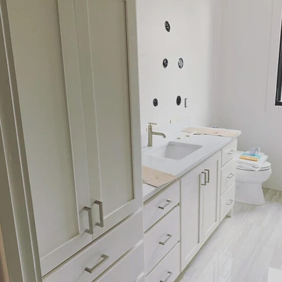 A modern bathroom with light-colored cabinets and drawers, a white countertop with a built-in sink, and black circular wall accents. Towels are stacked near the sink, and a toilet is visible by a window.