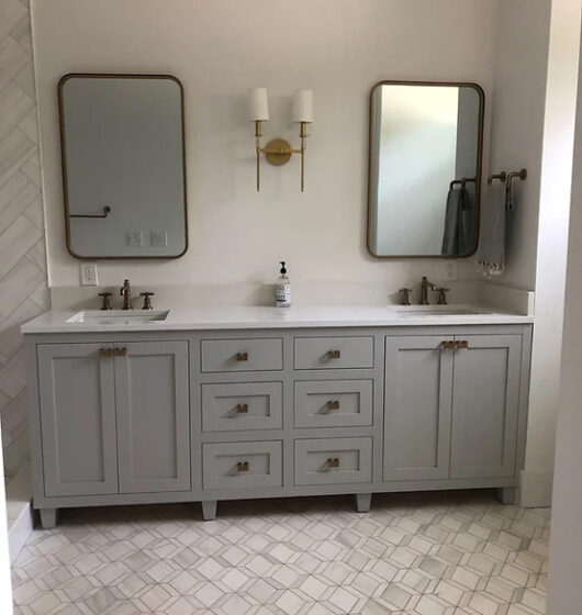 A modern bathroom with a double vanity featuring two rectangular mirrors, a central gold light fixture with two white shades, and a gray cabinet with six drawers. The countertop has two sinks and a soap dispenser, complemented by a tiled floor.