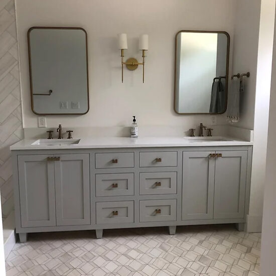 A modern bathroom with a double vanity featuring two rectangular mirrors, a central gold light fixture with two white shades, and a gray cabinet with six drawers. The countertop has two sinks and a soap dispenser, complemented by a tiled floor.