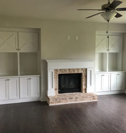 Living room with a white brick fireplace framed by built-in cabinets and shelves on both sides. The room features a dark hardwood floor and a ceiling fan with a light. The walls are painted a soft beige color.