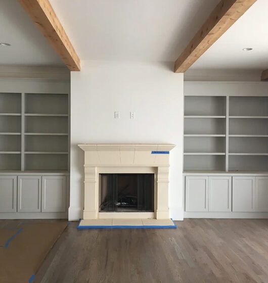 Living room with a light wooden floor, featuring a central fireplace with a beige mantel. Flanked by built-in shelves and cabinets on both sides. Exposed wooden beams on the ceiling add a rustic touch.