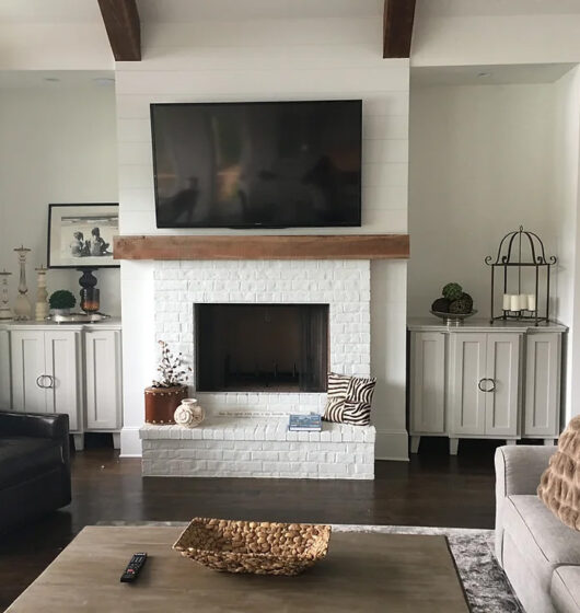 A cozy living room features a large TV mounted above a white brick fireplace. Beige cabinets flank the fireplace, and theres a black armchair and a gray sofa with a textured pillow. A coffee table with a woven basket sits on a gray rug.