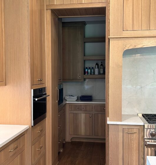 A cozy kitchen with light wood cabinetry and floors. The countertop is white, and theres a stainless steel oven. Shelves in a corner hold bottles and the backsplash is a light stone. The space appears modern and well-organized.