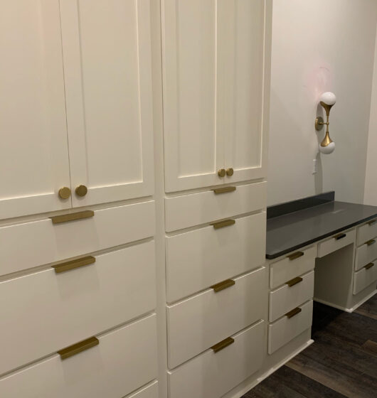 A modern interior with white cabinets featuring gold handles. To the right, theres a built-in desk with a dark countertop and a unique wall-mounted light fixture with two round bulbs. The floor is wooden with a dark finish.