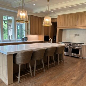 Spacious kitchen with wooden cabinets and a large center island featuring a white countertop and four woven stools. It includes stainless steel appliances, a double oven, and two pendant lights. Large windows offer a view of greenery outside.