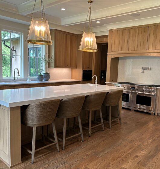 Spacious kitchen with wooden cabinets and a large center island featuring a white countertop and four woven stools. It includes stainless steel appliances, a double oven, and two pendant lights. Large windows offer a view of greenery outside.