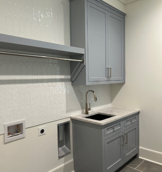 Laundry room with a light gray cabinet, granite countertop, and stainless steel sink. The wall features white hexagonal tiles. Below a rod for hanging clothes is a washing machine hookup and vent, set on a gray tiled floor.