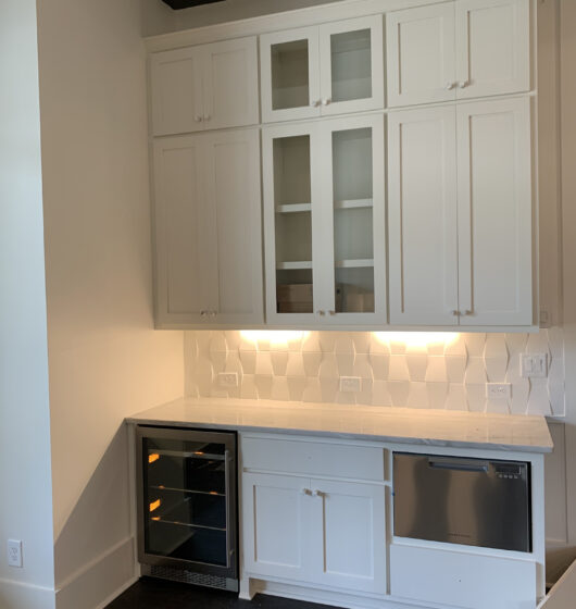 A modern kitchen with white cabinets, dark handles, and a geometric backsplash. Theres a small wine cooler to the left and a dishwasher to the right beneath the marble countertop. Soft under-cabinet lighting illuminates the counter.