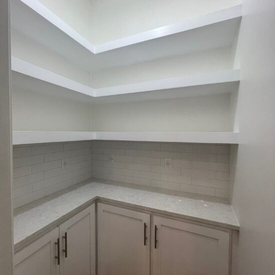 A clean, empty pantry with white shelves and light gray marble countertops. The walls are tiled with light gray subway tiles. The pantry features lower cabinets with silver handles on a wooden floor.