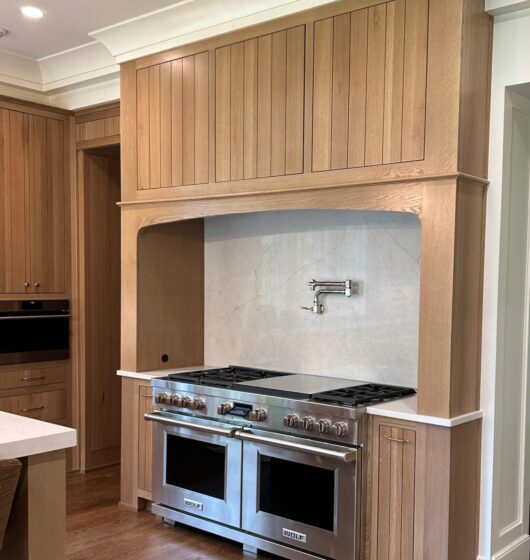 A modern kitchen features a double stainless steel stove with a sleek overhead pot filler. The stove is set in a wooden cabinet with vertical paneling, and the floor is made of dark wood. White walls and countertops complete the clean design.