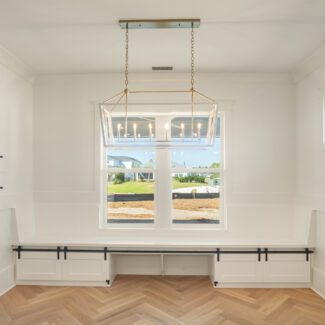 Bright, empty room with white walls and a large window. A geometric chandelier hangs from the ceiling. The floor features a herringbone wood pattern. Built-in bench seating runs along the walls below the window.