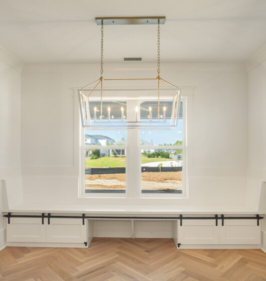 Bright, empty room with white walls and a large window. A geometric chandelier hangs from the ceiling. The floor features a herringbone wood pattern. Built-in bench seating runs along the walls below the window.