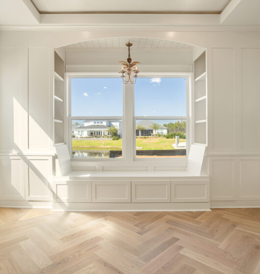 Bright room with large windows overlooking a waterfront. White paneled walls and a light wood herringbone floor enhance the airy atmosphere. A vintage-style chandelier hangs from the ceiling, illuminating the serene space.