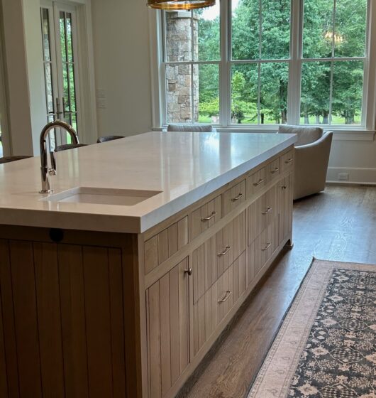 A modern kitchen features a large wooden island with a white countertop and a sink. Two pendant lights hang above. A rug lies on the wooden floor. Large windows offer a view of lush greenery outside.