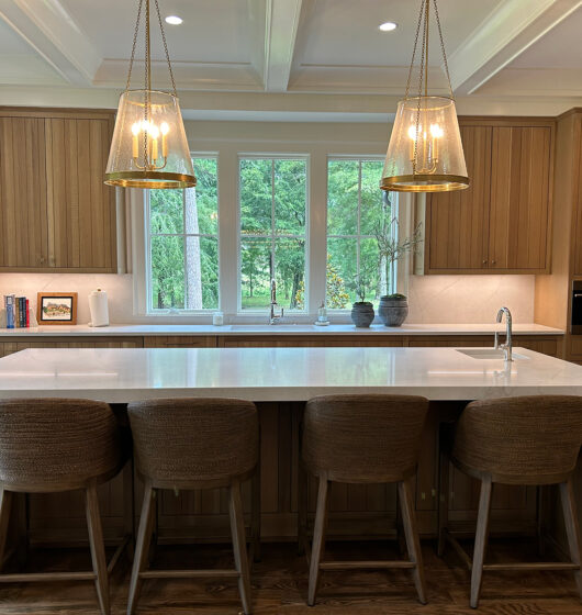 A modern kitchen with wooden cabinets and a large island featuring four woven stools. Two pendant lights hang above the island, and a window provides a view of greenery outside. The kitchen is equipped with built-in appliances.