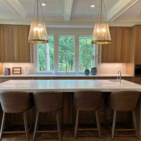 A modern kitchen with wooden cabinets and a large island featuring four woven stools. Two pendant lights hang above the island, and a window provides a view of greenery outside. The kitchen is equipped with built-in appliances.