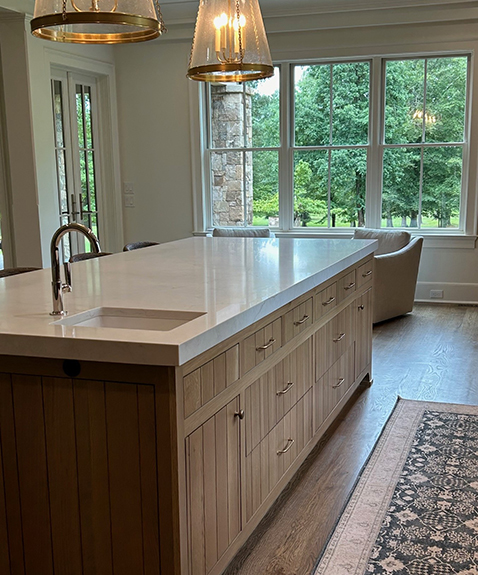 A modern kitchen with a large wooden island featuring a white countertop and built-in sink. Two pendant lights hang above. Large windows in the background offer a view of greenery. A patterned rug is partially visible on the wooden floor.