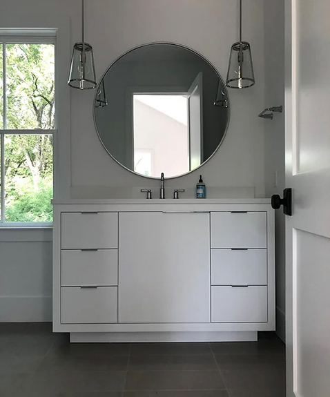 A minimalist bathroom with a round mirror above a white vanity cabinet. Two pendant lights hang on either side. A window on the left showcases greenery outside. A bottle sits on the sink, and the room features gray tile flooring.