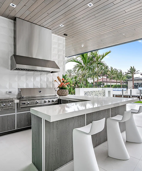 Modern outdoor kitchen with a sleek stainless steel grill and hood. The kitchen features a white countertop and stylish bar stools. Tropical plants and palm trees are visible in the background, with a partial view of a pool and waterfront.