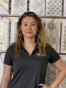 A woman with wavy, light brown hair wearing a dark gray polo stands in front of a display of decorative tiles with various patterns and shapes. She is smiling slightly and has one hand on her hip.