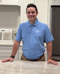 A man in a light blue polo shirt stands and smiles in a modern kitchen, resting his hands on a marble countertop. White cabinets and a stainless steel fridge are in the background.