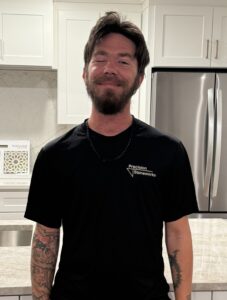 A man with tattoos on his arms, a beard, and dark hair stands smiling in a modern kitchen. He wears a black Precision Stoneworks t-shirt and a chain necklace. White cabinets and a stainless steel fridge are in the background.