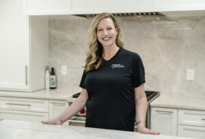 A smiling woman with long blonde hair stands in a modern kitchen, wearing a black shirt with a logo on the chest. She rests her hands on a marble countertop with white cabinets in the background.