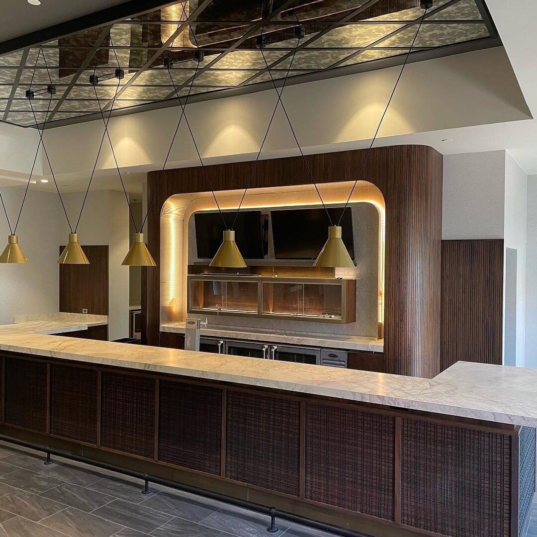 A modern, empty bar area with a marble countertop and wood paneling. Five gold pendant lights hang above the counter. The back wall features shelves and a TV, with warm recessed lighting outlining the structure.