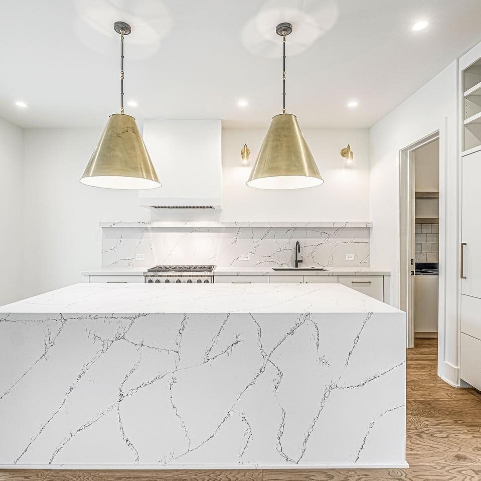 A modern kitchen with white marble countertops and island featuring gray veining. Two large gold pendant lights hang above the island. Stainless steel appliances are visible, with light wood flooring and a sliding glass door on the left.