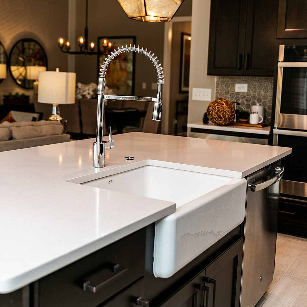 Modern kitchen interior featuring a sleek island with a white countertop and farmhouse sink. A chrome faucet is prominent. In the background, there are dark cabinets, a double oven, and a cozy living area with lamps and decorative mirrors.