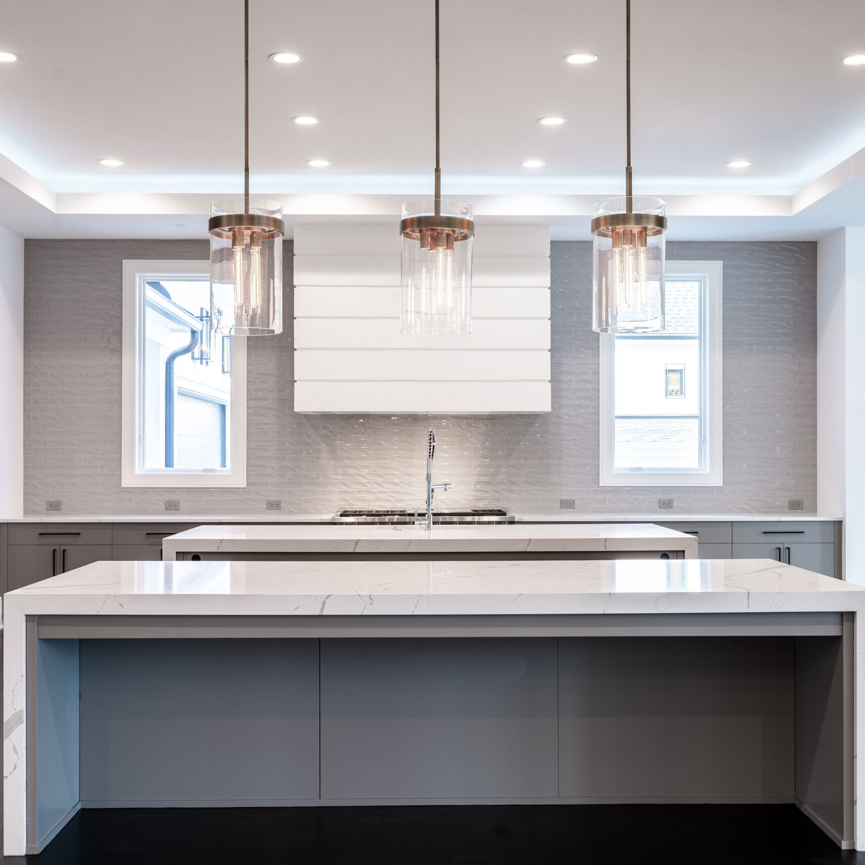 A modern kitchen with a large island and three pendant lights. The design features sleek cabinetry, stainless steel appliances, and a white marble countertop. Windows on either side allow natural light to illuminate the space.