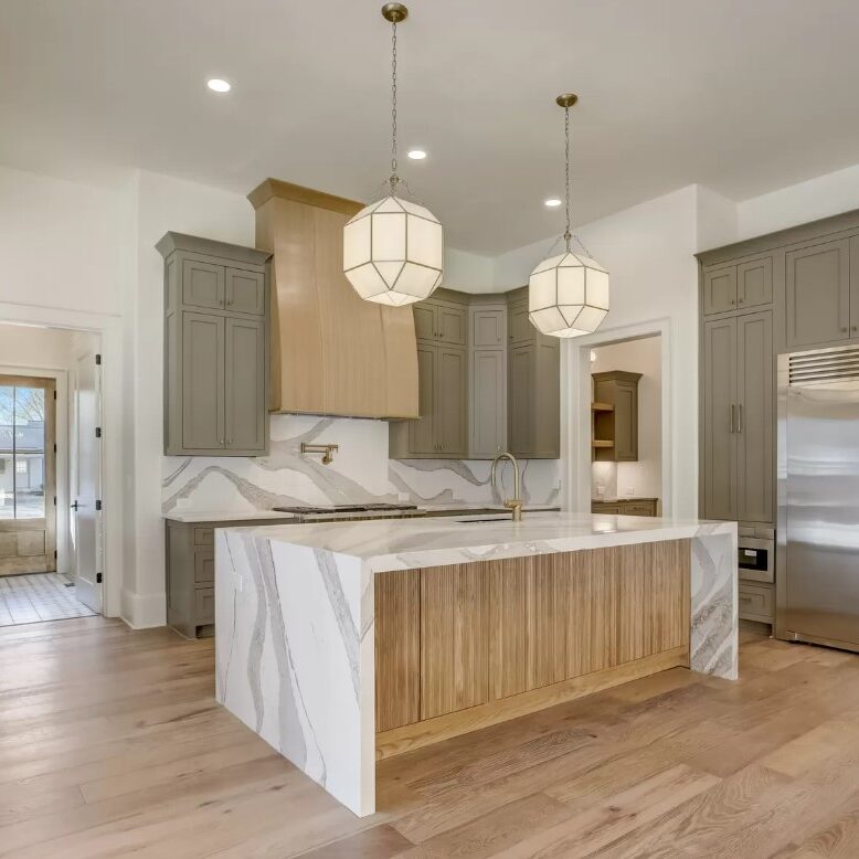 Modern kitchen with light wood flooring, marble countertops, and a large island featuring wooden accents. Gray cabinets and stainless steel appliances, including a double-door fridge. Two geometric pendant lights hang over the island.