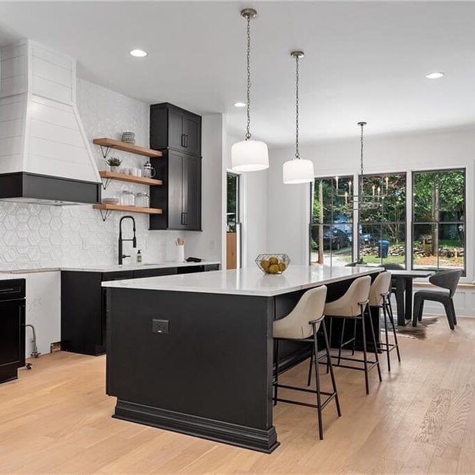 Modern kitchen with a large white island countertop, black base, four bar stools, and pendant lights. White hexagonal tile backsplash, black and wood cabinets, a black sink, and floating shelves. Adjacent dining area with large windows and a table.