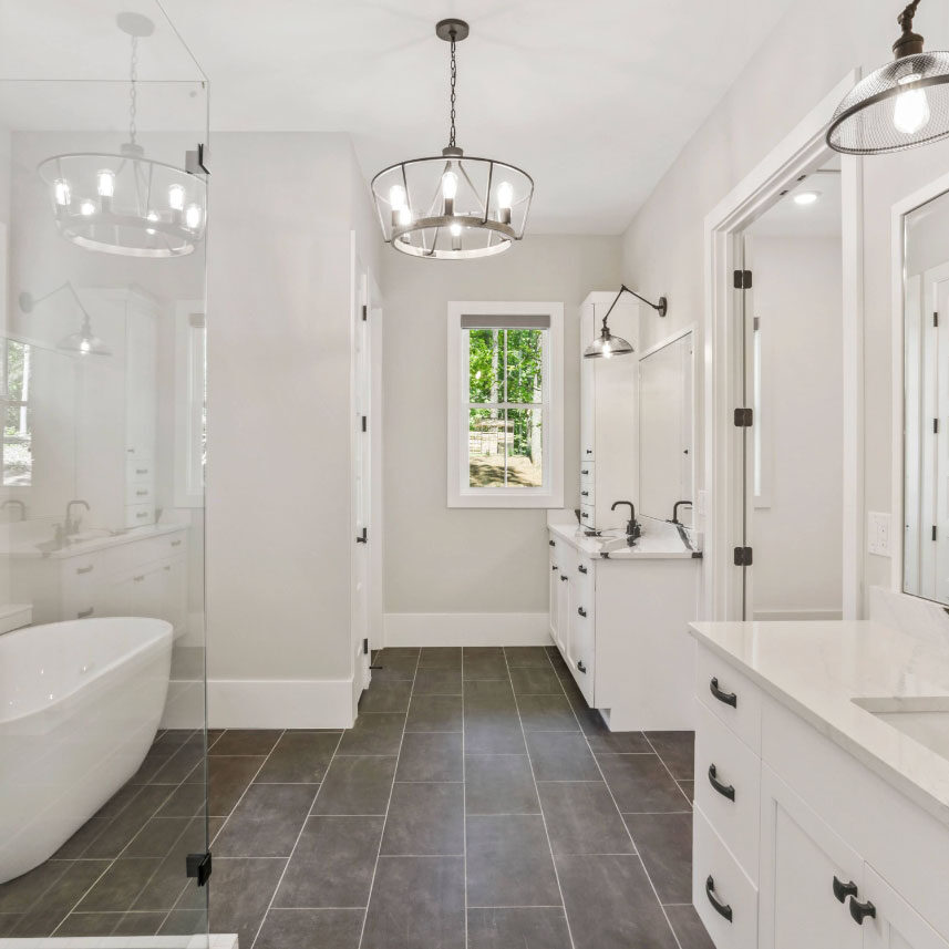 Modern bathroom with a freestanding white bathtub beside a glass shower enclosure. Double sink vanity with marble countertops and black faucets. Pendant lights hang from the ceiling. Large mirror and window bring in natural light, accentuating gray tiles.